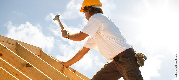 Dachdecker mit gelbem Schutzhelm beim Befestigen von Konstruktionsvollholz (KVH) auf einem Dachstuhl unter strahlendem Himmel.